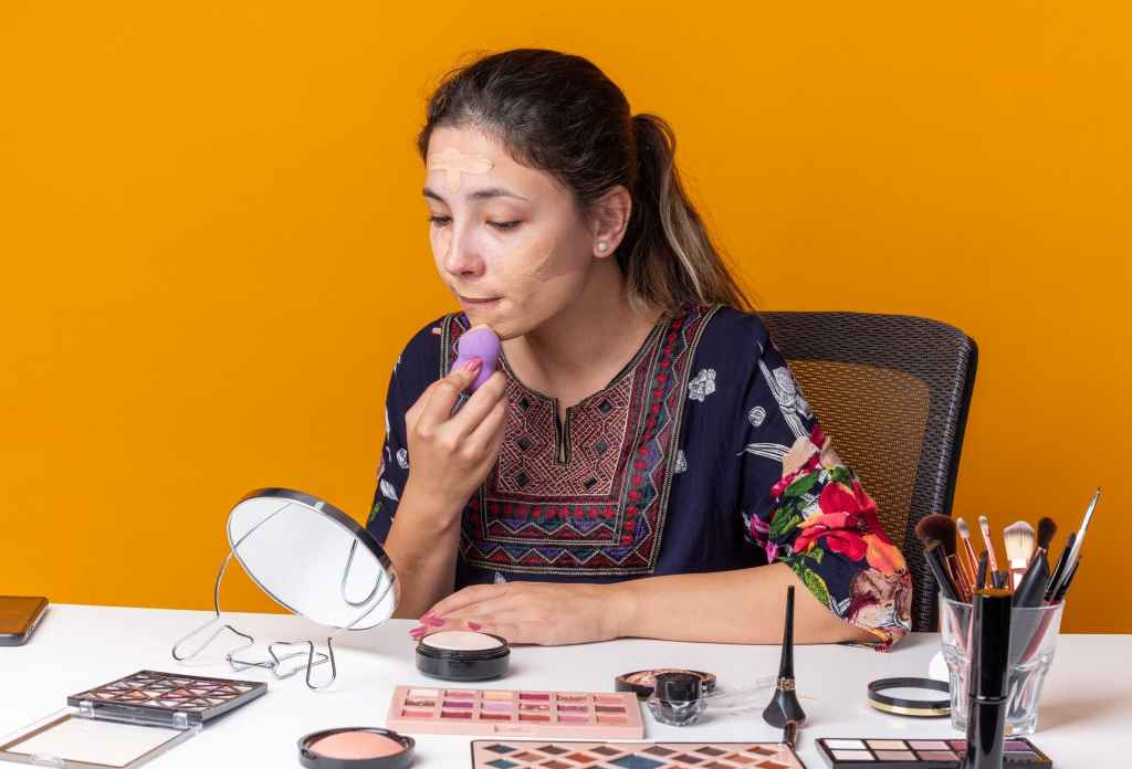 girl sitting at table with makeup tools applying foundation